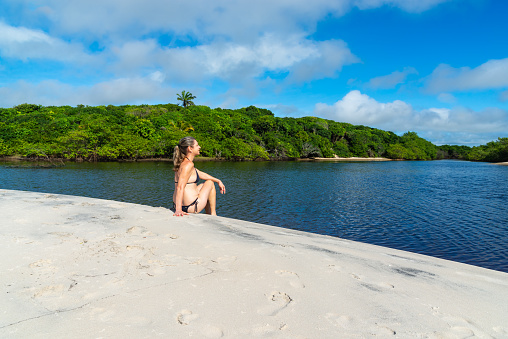 A woman sitting by the river against the forest in the background. Guaibim beach in the city of Valenca, Bahia, Brazil.