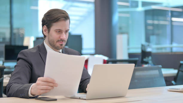istock Businessman with Laptop Reading Documents in Office 1421432906