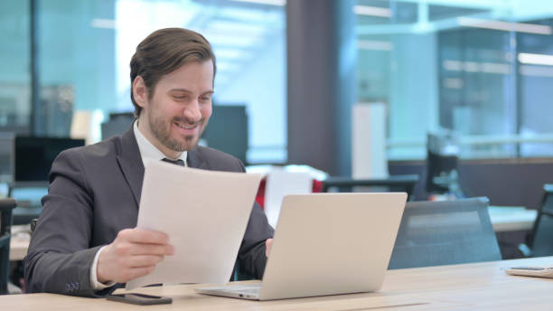 istock Businessman with Laptop Celebrating Success while Reading Documents 1421433136