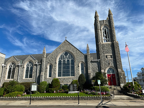 horizontal image of a quaint little white country church with a steeple sitting in a green meadow surrounded by trees under a beautiful blue sky with white clouds floating by in the summer time.