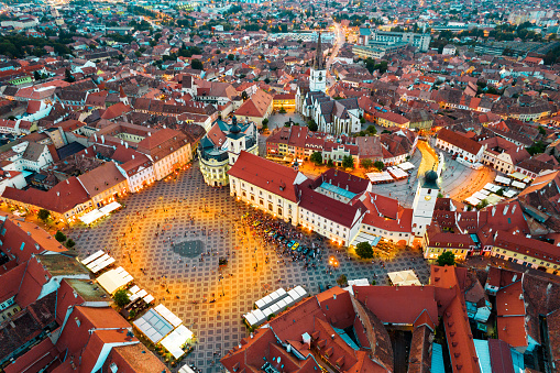 Aerial drone view of the Historic Centre of Sibiu at evening, Romania. The Great Square with Sibiu Lutheran Cathedral and old buildings around, narrow streets, people, illumination