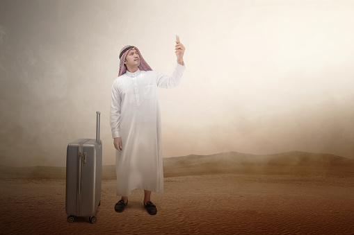 Arab man wearing keffiyeh looking at mobile phone with a suitcase for traveling on the desert