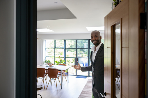 Welcoming real estate agent opening the door of a house for sale and looking at the camera smiling