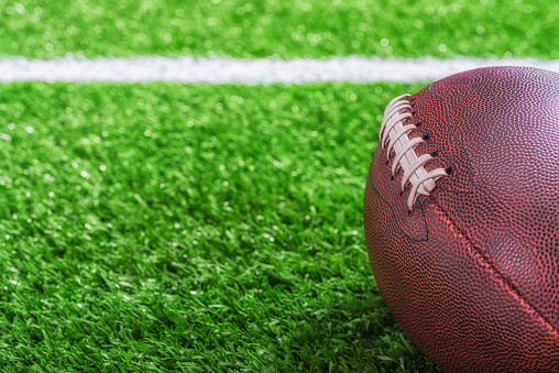 A low angle close-up view of an American Football sitting on the turf with a white yard line in the background.