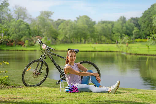 asian female biker sitting beside bicycle by lake in park relaxing after cycling for exercise for wellbeing life during summer