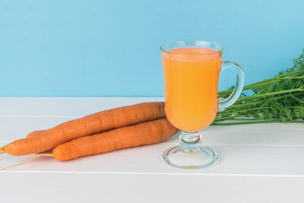 a glass of juice and fresh carrots on a white table on a blue background. - carotene imagens e fotografias de stock