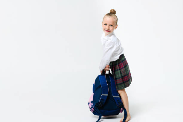 Cute girl in a school uniform with a big and heavy backpack on a white background. Girl lifts a huge school backpack.