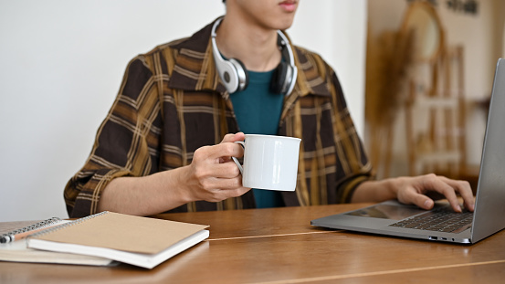 A handsome young Asian male college student or freelancer with wireless headphones, working on his project on laptop while sipping a morning coffee. cropped image