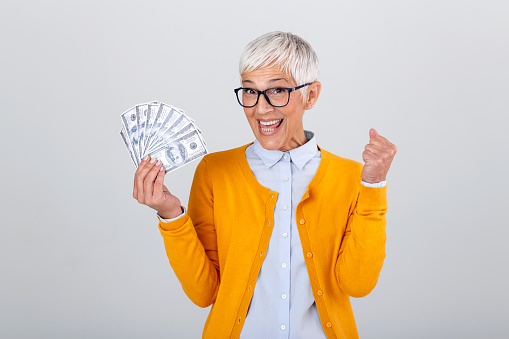 Beautiful senior lady holding a bunch of $100 bills with surprised facial expression. Mature woman winning money. Finances and people concept, senior woman holding hundreds of dollar money banknotes