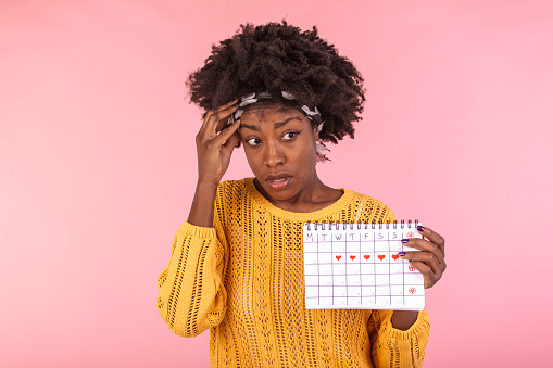 Young African American woman holding menstruation calendar over isolated background with surprise face. Woman holding menstruation calendar with confused face, afraid and excited with fear expression