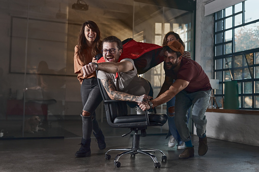 Playful creative people having fun on a break at casual office while pushing their colleague on a chair who is pretending to be a superman.