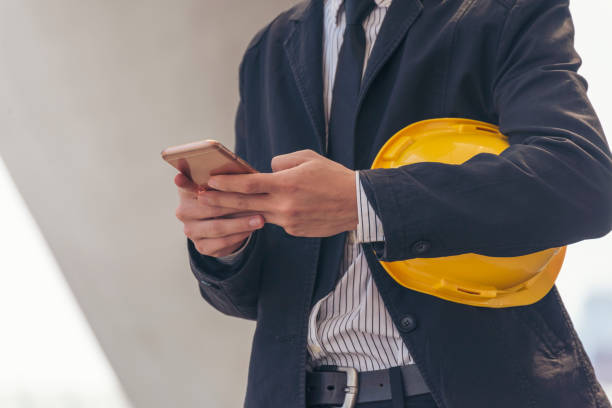 istock Construction engineer holding yellow hard hat holding smartphone. Safety helmet Holding Yellow hardhat using smartphone to communicate on Construction Site. Hardhat Protect Civil Construction concept 1421513214