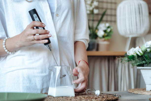 mélanger le lait dans un pot en verre en gros plan. femelle fabriquant du lait mousseux avec un mélangeur portatif. femme au foyer vêtue de vêtements domestiques à l’aide de fouets mélangeurs cuisant des aliments dans la cuisine. concept de cuisson - foamed milk photos et images de collection