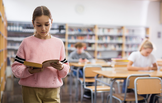 Preteen girl standing alone near bookcase in library browsing textbooks