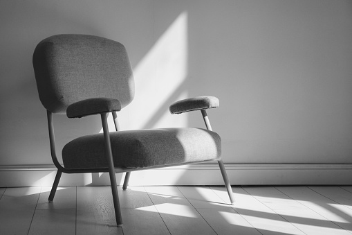 Black and white image of stylish armchair in interior domestic room with sunlight from window. Home composition of cozy living room with chair of shadow background. Copy text space