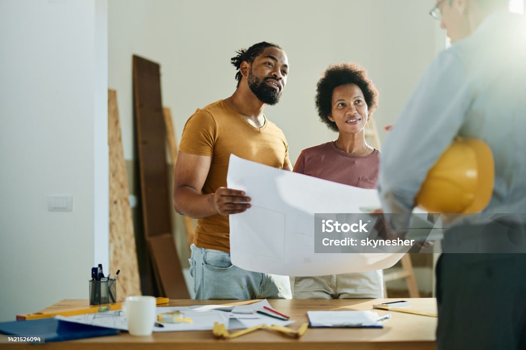 Happy black couple talking to real estate agent in the apartment. Happy African American couple analyzing blueprints while communicating with a building contractor in the apartment. Renovation Stock Photo