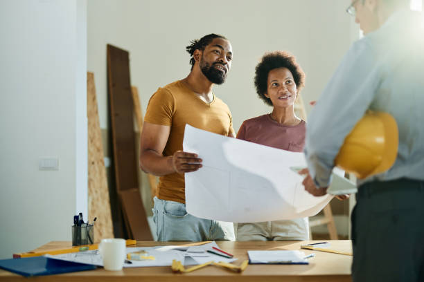 Happy black couple talking to real estate agent in the apartment. Happy African American couple analyzing blueprints while communicating with a building contractor in the apartment. building contractor stock pictures, royalty-free photos & images