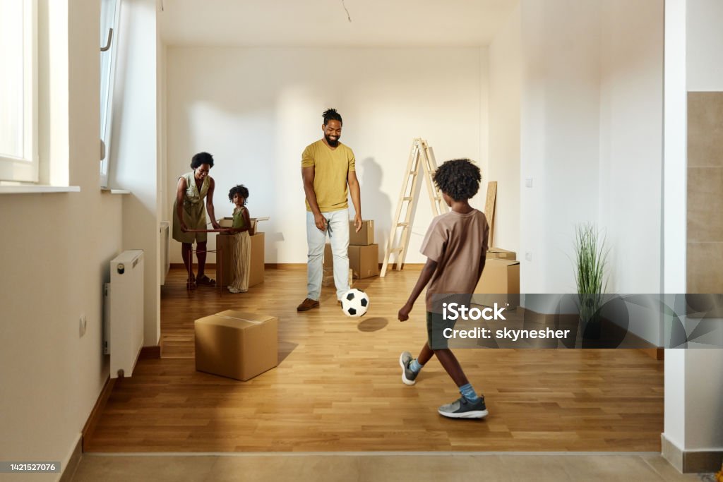 Happy black family having fun while unpacking in new apartment. Happy African American father and son playing soccer while mother and daughter are unpacking their belongings after moving into a new apartment. African Ethnicity Stock Photo