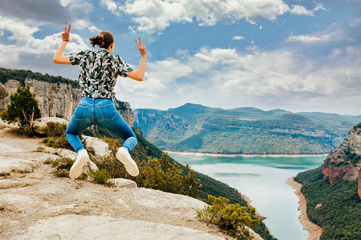 Rear view portrait of young cheerful woman jumps in mid air and does victory sign on top of mountain lake. Happy female wears floral shirt and ponytail. Adventure, travel, holiday concept.