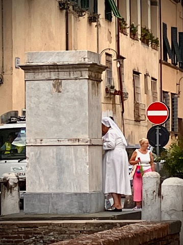 Catolic Nun dressed in a white gown getting water by the fountain the City of Lucca Italy on a cloudy autumn afternoon, 6rd September 2022. Lucca is a city on the Serchio river in Italy’s Tuscany region. It’s known for the well-preserved Renaissance walls encircling its historic city center and its cobblestone streets. Broad, tree-lined pathways along the tops of these massive 16th- and 17th-century ramparts are popular for strolling and cycling. Casa di Puccini, where the great opera composer was born, is now a house museum.