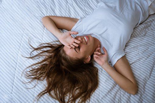 Good morning. Leisure of the day off. Well-being for health. Happy relaxed brunette woman stretching out a smile while lying in bed in home bedroom covering her face with hands for fun