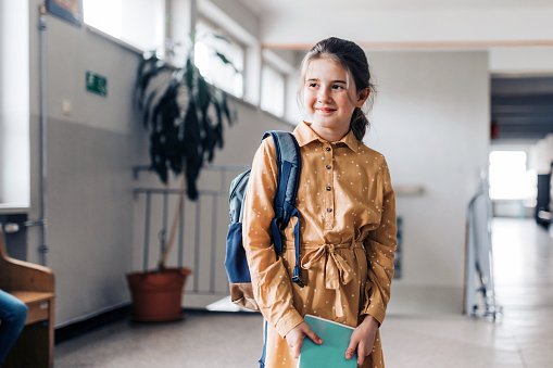 portraits of happy smiling elementary schoolgirl holding workbook  in corridor