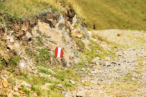 Red and white trail sign (trail marker) painted on a stone alongside a hiking footpath in Austrian Alps. Feistritz an der Gail municipality, Carinthia, Carnic Alps, Italy-Austria border, Europe.