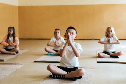 elementary school children sitting cross-legged on the floor of gym and meditating in yoga lesson