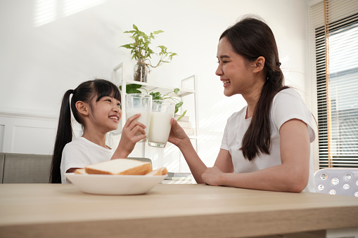 A Healthy Asian Thai family, little daughter, and young mother drink fresh white milk in glass and bread joy together at a dining table in morning, wellness nutrition home breakfast meal lifestyle.