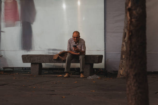 El Espinar, Spain - September 3, 2022: A lonely elderly man watches a smartphone while sitting on a stone bench on a city square.