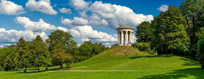 The round temple or Monopteros temple in the English garden in Munich in panoramic view with meadows and deciduous trees in summer weather with loosened clouds