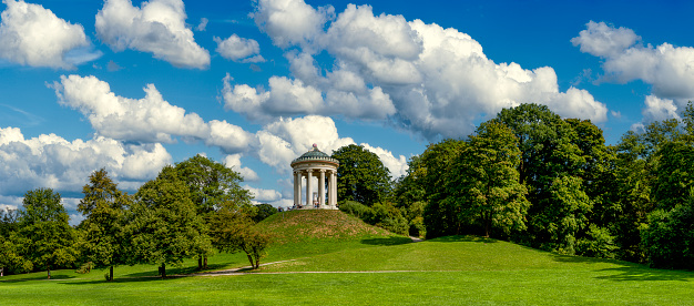 The round temple or Monopteros temple in the English garden in Munich in panoramic view with meadows and deciduous trees in summer weather with loosened clouds