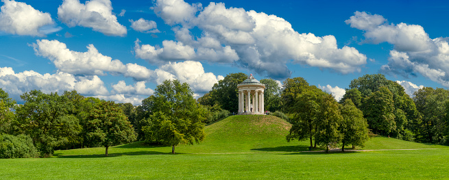 The round temple or Monopteros temple in the English garden in Munich in panoramic view with meadows and deciduous trees in summer weather with loosened clouds