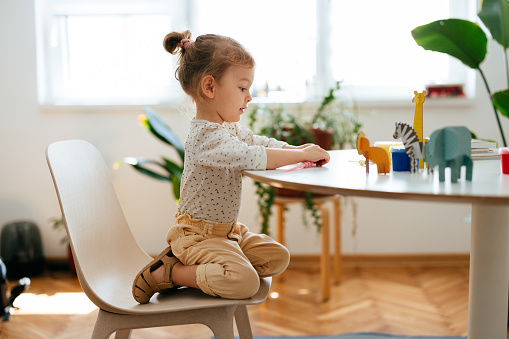 Cute little girl is crouching on the chair playing with paper animals that are on the table.