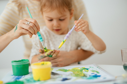 Portrait of a cute little girl sitting in her anonymous mother's lap looking interested in what her mom is doing while mixing the paint with two brushes and painting it on her hand.