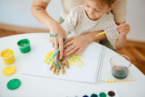 Unrecognizable woman is sitting with her daughter in her lap. They are having fun with paint while leaving coloured handprints on the piece of paper in front of them.