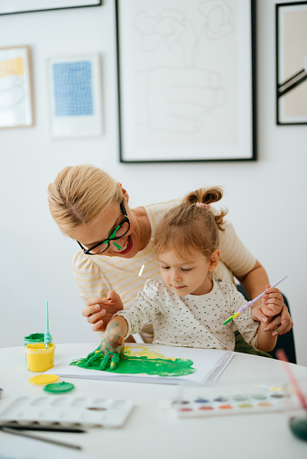 Portrait of a happy beautiful woman sitting with her daughter at the table and having fun while playing with paint. They are excited to paint the girl's palm and leave handprints on paper. Both of them are smiling. The woman has paint on her glasses.