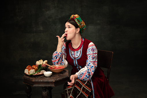 Pampushkas, borscht and salo. Emotional young woman wearing national folk Ukrainian attire isolated over dark retro background. Human emotions, history, ad, fashion, beauty, eras comparison concept.
