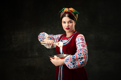 Creative portrait of beautiful Ukrainian woman wearing traditional folk costume isolated over dark vintage background. Fashion, beauty, cultural heritage concept. Human emotions