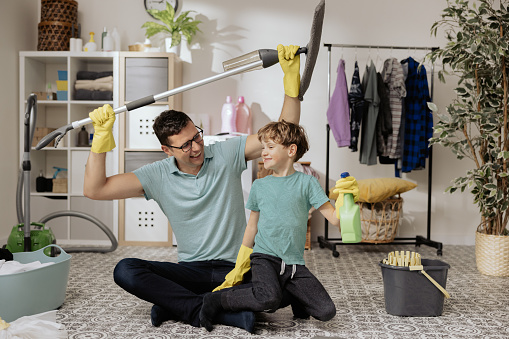 Cheerful happy man holds son on lap, child helps dad clean house, uses liquid detergent, guy raises floor mop above his head, they fool around, fun.