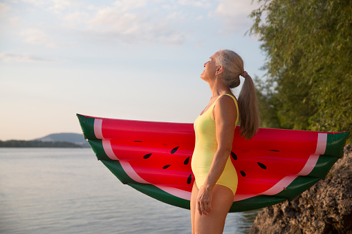Summer lifestyle portrait senior woman in swimwear with air mattress looks like watermelon on the seashore. Enjoying the little things. spends time in nature in summer