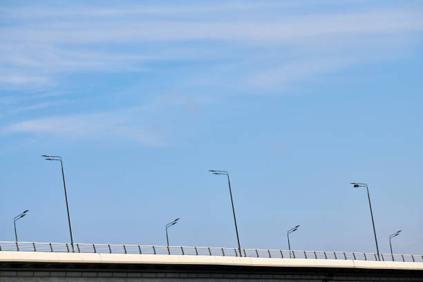 Concrete city bridge over river with street lampposts, angle view with blue sky copy space background. Minimalistic bridge with automobile highway and wireless lamp posts, modern city infrastructure