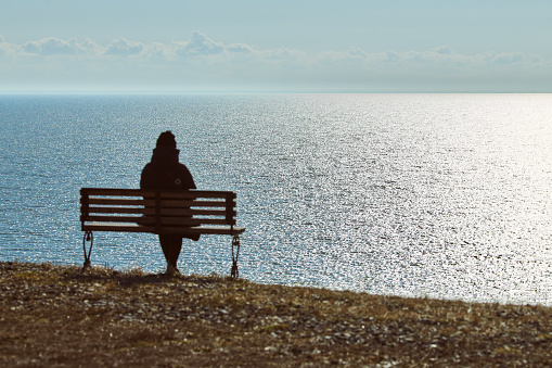 Single girl in a black jacket and hat sitting on bench at cliff at front of sea, peaceful and quiet place for thinking alone, loneliness and loss of loved one concept. Pacifying view of marine horizon
