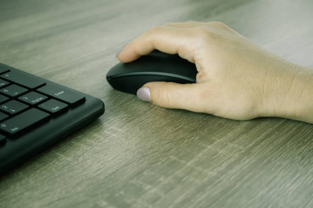 istock A woman's hand works with a mouse and keyboard manipulator. Caucasian Woman using a black wireless computer mouse on wooden background. Female hand with modern computer mouse 1421577352