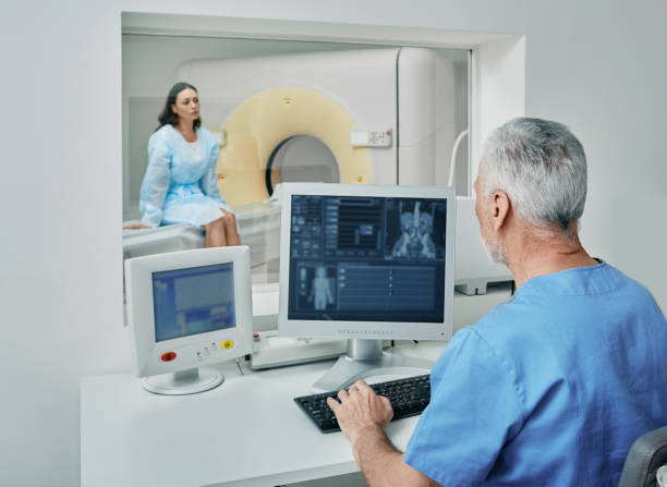 radiologe vor ct-scan des weiblichen körpers mit ct-scanner aus dem kontrollraum hinter dem schutzfenster. computertomographie - medical equipment x ray cancer oncology stock-fotos und bilder