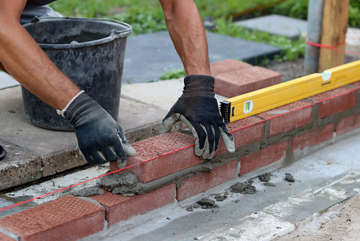 Male hands in protective gloves holding red bricks. Construction worker builds masonry.
