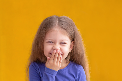Portrait of laughing caucasian little girl child kid of 5 years looking at camera covering mouth by hand giggle on yellow background