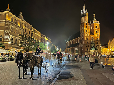 St Mary's Basilica and horse drawn carriages in Rynek Główny medieval main square night lights, Krakow, Poland. Krakow city in Poland was originally the capital of the country until 1956 but is now best known for its well-preserved medieval centre with its period architecture and spacious Rynek Glówny market square as well as the Jewish quarter and ghetto areas.