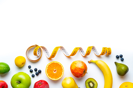 Overhead view of a composition of healthy fruits and a yellow tape measure shot on white background. The composition is at the bottom of an horizontal frame making a border and leaving copy space.  High resolution 42Mp studio digital capture taken with SONY A7rII and Zeiss Batis 40mm F2.0 CF lens