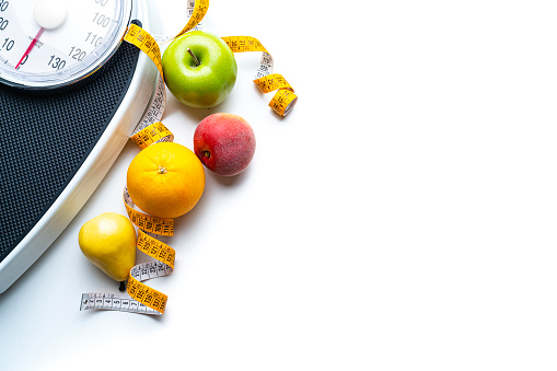 Overhead view of a composition of healthy fruits, weight scale, tape measure and dumbbells shot on white background. The composition is at the top of an horizontal frame making a border and leaving copy space.  High resolution 42Mp studio digital capture taken with SONY A7rII and Zeiss Batis 40mm F2.0 CF lens